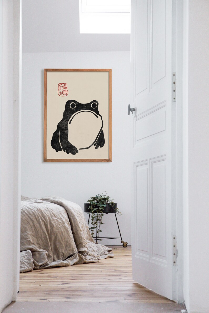 Large framed Japanese frog illustration hangs in a bright minimalist bedroom with natural linens and soft wooden flooring. The quirky frog and red seal detail bring a touch of humor and refined calm, blending seamlessly with Japandi and zen decor.