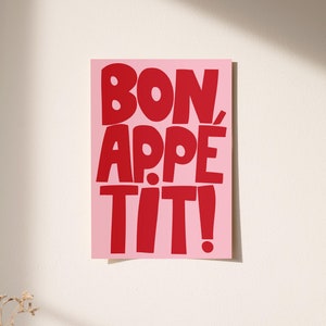 Bold red typography reading "Bon Appétit" on a soft pink background, displayed unframed against a sunlit cream wall. The playful and vibrant lettering casts a subtle shadow, creating a lively and cheerful atmosphere ideal for kitchen or dining room decor. Warm light and minimal styling highlight the graphic print’s fun, retro-inspired charm.