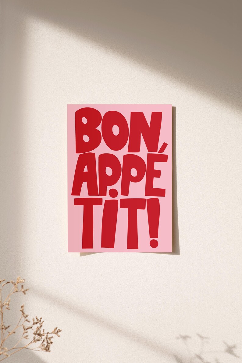 Bold red typography reading "Bon Appétit" on a soft pink background, displayed unframed against a sunlit cream wall. The playful and vibrant lettering casts a subtle shadow, creating a lively and cheerful atmosphere ideal for kitchen or dining room decor. Warm light and minimal styling highlight the graphic print’s fun, retro-inspired charm.