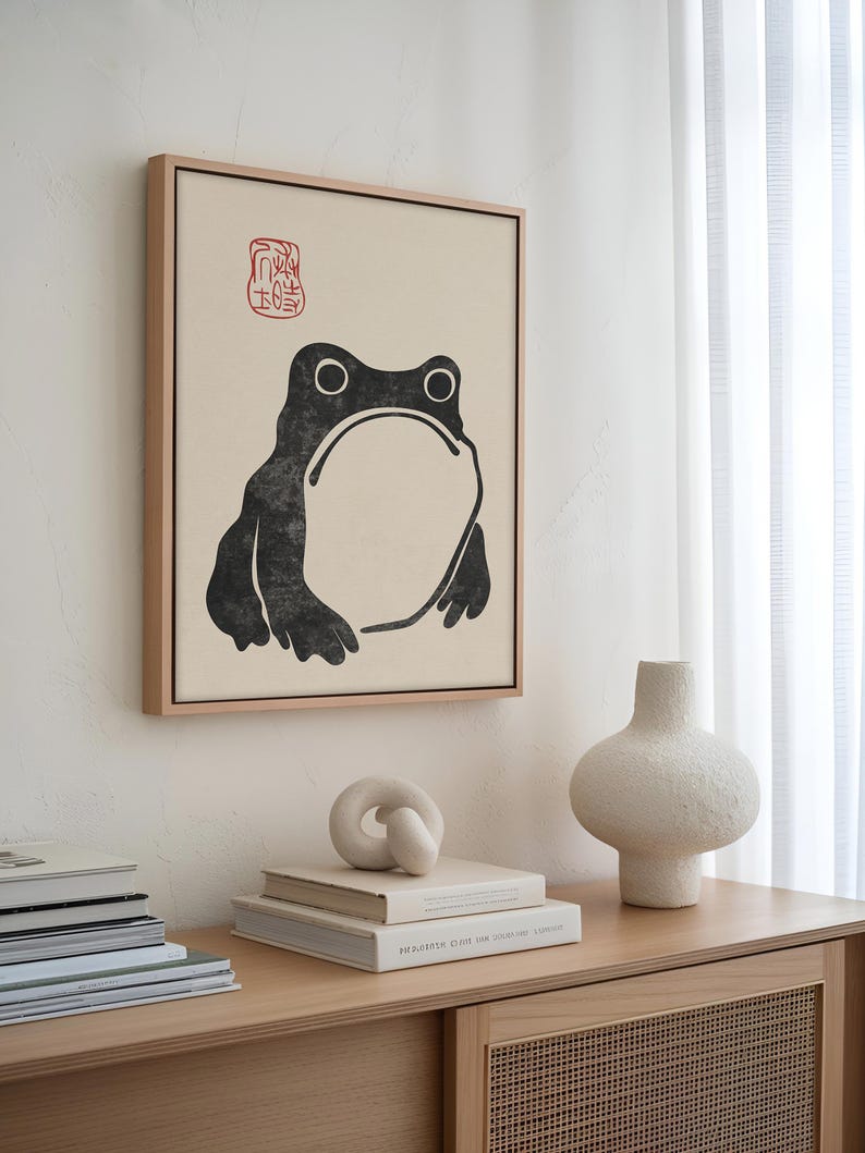 Minimal frog artwork by Matsumoto Hoji displayed above a wooden sideboard styled with sculptural decor and stacked books. The black and cream color palette, paired with the delicate red stamp, creates a clean focal point for Japandi or minimalist spaces.