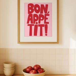 Framed "Bon Appétit" typography print with bold red letters on a pink background, hanging on a minimalist kitchen wall above a wooden counter and a bowl of red apples, creating a cheerful and inviting dining space with retro color contrast and modern decor appeal.