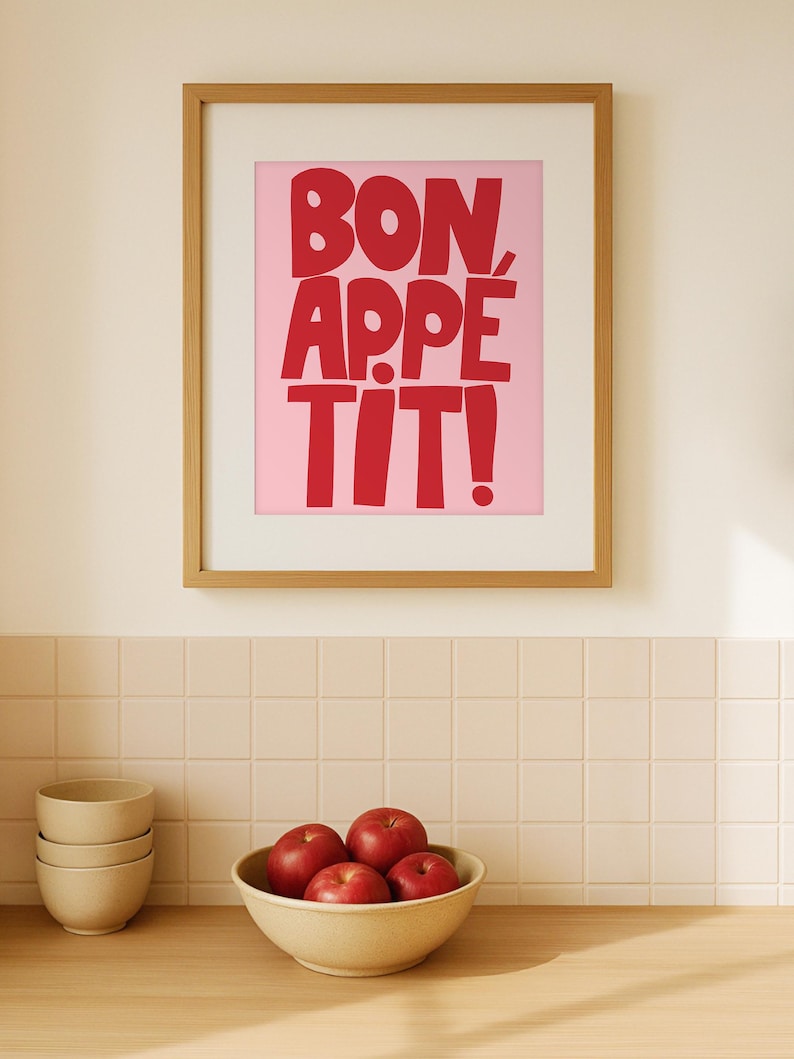 Framed "Bon Appétit" typography print with bold red letters on a pink background, hanging on a minimalist kitchen wall above a wooden counter and a bowl of red apples, creating a cheerful and inviting dining space with retro color contrast and modern decor appeal.