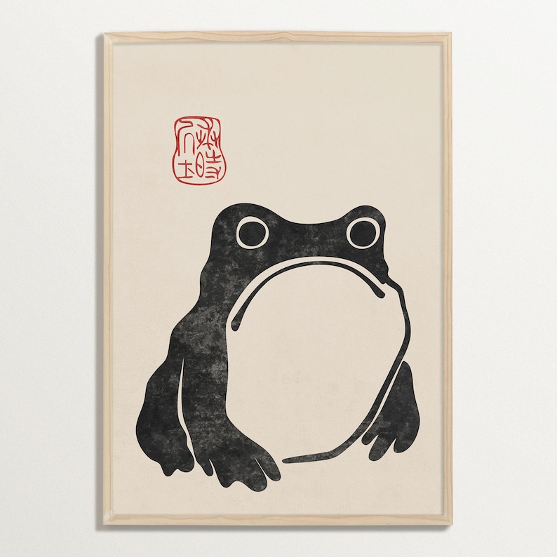 Framed minimal Japanese frog illustration on a soft cream background, featuring bold black ink lines and a red seal in the top left. The whimsical expression and clean design by Matsumoto Hoji evoke zen simplicity and traditional woodblock charm.