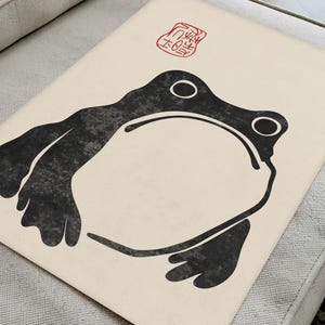 Flat-lay of Matsumoto Hoji’s unimpressed frog print on a light upholstered surface. The organic textures of the paper and furniture enhance the bold monochrome frog illustration, perfect for lovers of traditional Japanese ink art and contemporary minimalism.