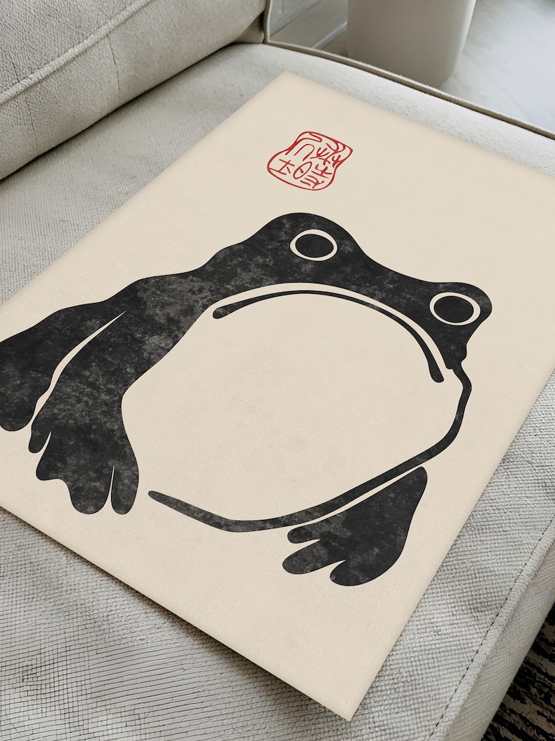Flat-lay of Matsumoto Hoji’s unimpressed frog print on a light upholstered surface. The organic textures of the paper and furniture enhance the bold monochrome frog illustration, perfect for lovers of traditional Japanese ink art and contemporary minimalism.