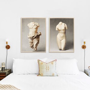 May include: Two framed prints of white marble sculptures of torsos, one male and one female, against a white wall with a bed in the foreground.