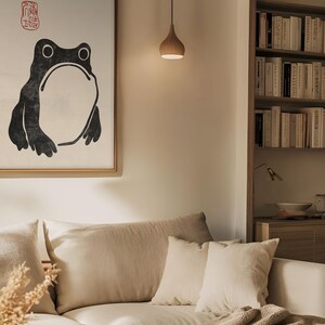 Japanese frog print by Matsumoto Hoji framed above a cozy beige sectional in a softly lit, earthy-toned living room. The expressive ink illustration and red seal stamp stand out in a calm, neutral setting, adding character and wabi-sabi charm to modern interiors.