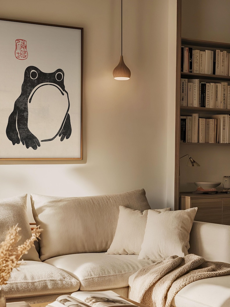 Japanese frog print by Matsumoto Hoji framed above a cozy beige sectional in a softly lit, earthy-toned living room. The expressive ink illustration and red seal stamp stand out in a calm, neutral setting, adding character and wabi-sabi charm to modern interiors.