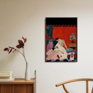 Framed George Barbier Art Deco print hanging above a wooden cabinet in a modern minimalist room. The erotic vintage illustration shows two nude women in an intimate embrace, with rich red tones and classical motifs, ideal for sophisticated boudoir or eclectic art spaces.
