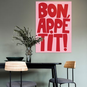 Large-format "Bon Appétit" poster in vivid red and pink colors displayed above a black dining table with modern chairs and minimalist greenery, blending pop-art typography with Scandinavian interior styling.