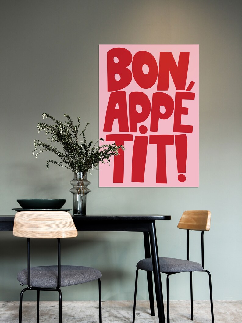 Large-format "Bon Appétit" poster in vivid red and pink colors displayed above a black dining table with modern chairs and minimalist greenery, blending pop-art typography with Scandinavian interior styling.