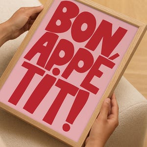 Close-up of hands holding a framed "Bon Appétit" poster with playful red block lettering on a bubblegum pink background, showcasing the compact size and bold visual impact of the kitchen-themed wall art in a casual home setting.