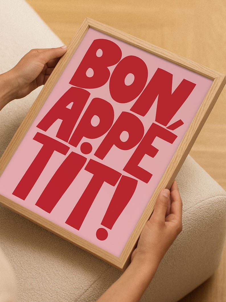 Close-up of hands holding a framed "Bon Appétit" poster with playful red block lettering on a bubblegum pink background, showcasing the compact size and bold visual impact of the kitchen-themed wall art in a casual home setting.