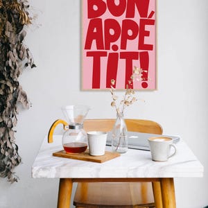 Framed "Bon Appétit" print in bold red on pink, hanging above a cozy breakfast nook with a marble table, coffee cups, and natural wood furniture, enhancing the space with a vibrant and cheerful graphic element perfect for kitchen decor.