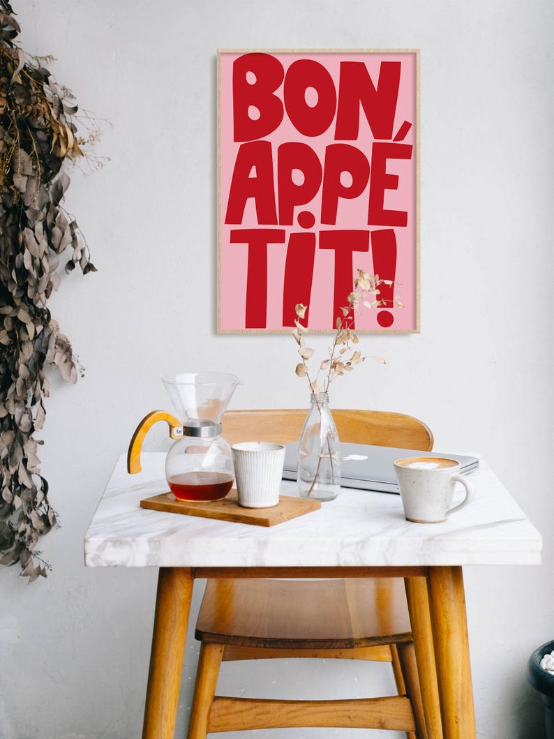 Framed "Bon Appétit" print in bold red on pink, hanging above a cozy breakfast nook with a marble table, coffee cups, and natural wood furniture, enhancing the space with a vibrant and cheerful graphic element perfect for kitchen decor.