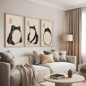 Minimalist Japanese ink painting of a serious frog holding a tea whisk, printed on cream-colored textured paper. The black brushstrokes create a striking wabi-sabi aesthetic. Displayed above a sofa with neutral decor, the artwork adds a touch of Zen charm and playful character to the space.