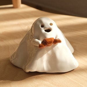 Handmade ceramic incense burner or candle holder, shaped like a ghost holding a croissant with both hands. It has holes in the eyes and mouth. So, you can see incense smoke or candlelight coming out these holes.