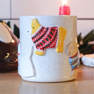 May include: White ceramic mug with a festive design featuring a yellow dog wearing a red and white sweater, a white dog wearing a blue and white sweater, and a yellow dog wearing a red and white sweater. The mug is sitting on a wooden surface with a lit candle in the background.