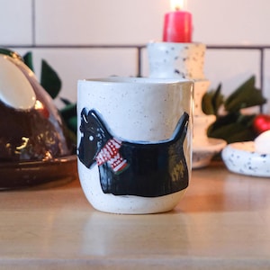 May include: A white ceramic mug with a black and white silhouette of a dog wearing a red and white scarf. The mug is sitting on a wooden surface with a Christmas pudding and candles in the background.