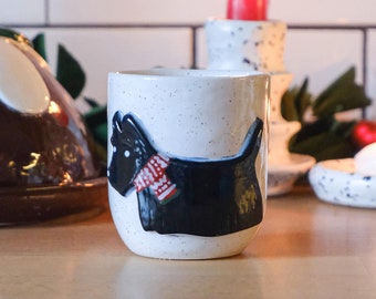Puede incluir: Una taza de cerámica blanca con una silueta negra y blanca de un perro que lleva una bufanda roja y blanca. La taza está sobre una superficie de madera con un pudín de Navidad y velas en el fondo.