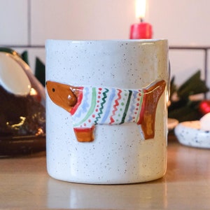 May include: White ceramic mug with a brown dog wearing a colorful sweater design. The mug is on a wooden surface.