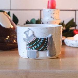 May include: A white ceramic mug with a gray dog wearing a green and red sweater design. The mug is sitting on a wooden surface with a Christmas pudding and candles in the background.