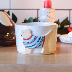 May include: A ceramic candle holder with a light beige speckled finish. It features a 3D dog wearing a blue, red, and white sweater. The candle holder is on a wooden surface, with a lit red candle in the background.