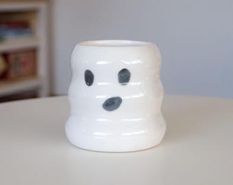 May include: A white ceramic ghost-shaped mug with a wide opening. The mug has a rounded body with horizontal ridges and a painted face with two black eyes and a gray mouth. The mug is sitting on a white table.