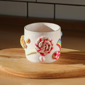 Puede incluir: Una taza de cerámica blanca con un esmalte moteado, decorada con formas coloridas que parecen caramelos, incluyendo un remolino rojo y blanco, un arcoíris y un remolino amarillo.