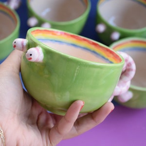 Può includere: Ciotole in ceramica verde con un design arcobaleno sul bordo e manici rosa a forma di verme. Le ciotole sono esposte su uno sfondo viola e blu.