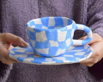 May include: A blue and white checkered ceramic teacup and saucer. The cup and saucer have a hand-painted design.