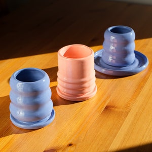 Pode incluir: Três vasos de cerâmica, dois de cor azul claro e um de cor rosa claro. Os vasos têm um design texturizado e ondulado e estão sobre uma superfície de madeira.