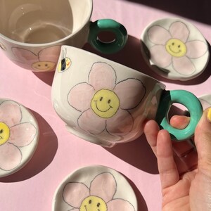 Può includere: Set di quattro tazze da tè e piattini in ceramica con un design a fiore rosa e un viso sorridente. Le tazze hanno un manico verde.