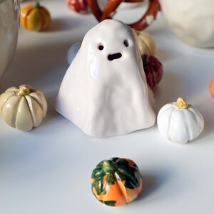 May include: A white ceramic ghost figurine with a simple face, surrounded by small ceramic pumpkins in various colors, including orange, green, and white.