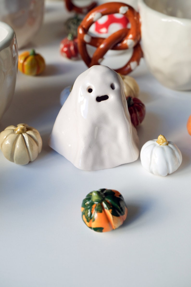 May include: A white ceramic ghost figurine with a simple face, surrounded by small ceramic pumpkins in various colors, including orange, green, and white.