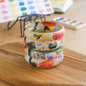 Może przedstawiać: Stos trzech ceramicznych kubków z kolorowym abstrakcyjnym wzorem. Kubki są ułożone na drewnianej desce do krojenia. Kubki są pomalowane plamami czerwonej, żółtej, niebieskiej, zielonej i czarnej farby.