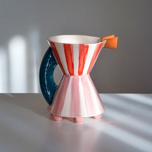 Op de afbeelding: Een keramische kan met een gestreept ontwerp in roze en rood. De kan heeft een teal handvat en een kleine oranje tuit. De kan heeft een brede basis met vier kleine voetjes.