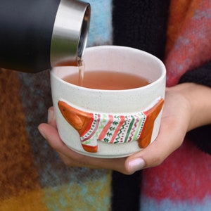 May include: A white ceramic mug with a brown dog wearing a colorful sweater design. The mug is filled with a light brown liquid and a black thermos is pouring the liquid into the mug.