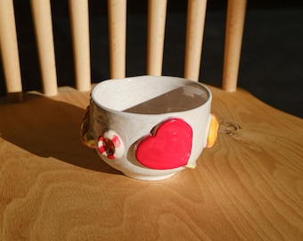May include: A white ceramic bowl with a speckled glaze, decorated with three-dimensional candy-like shapes. The bowl features a red heart, a yellow swirl, and a red and white candy cane.