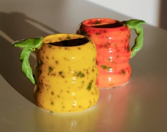 May include: Two ceramic mugs with green leaf handles. One mug is yellow with green speckles and the other is orange with red speckles.