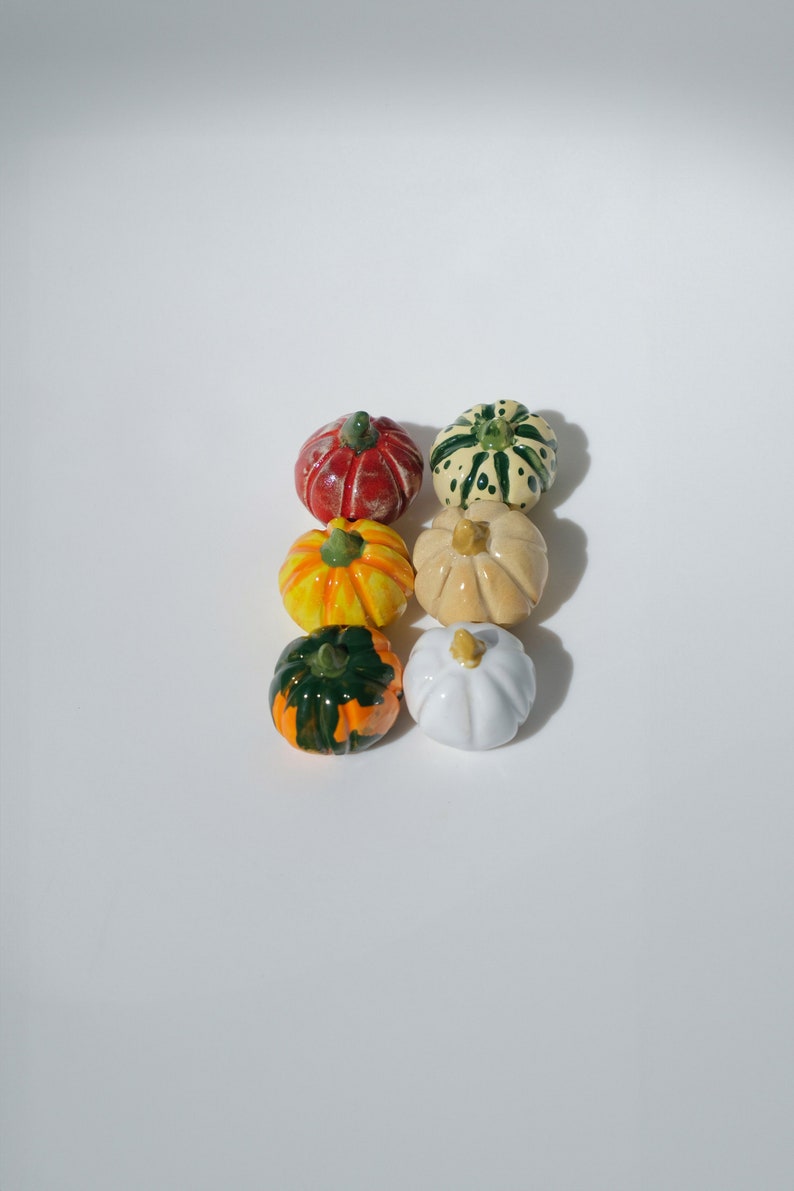 May include: Six miniature ceramic pumpkins in various colors and patterns, including green, white, yellow, orange, and red. The pumpkins are arranged in a row on a white surface.