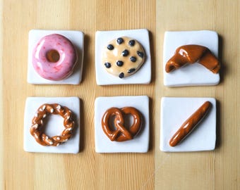 May include: A collection of six ceramic food-themed coasters. The set includes a pink glazed donut, a chocolate chip cookie, a croissant, a pretzel, a soft pretzel, and a baguette. Each coaster is square and white.