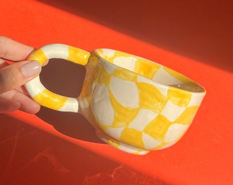 May include: A yellow and white ceramic mug with a checkered pattern. The mug has a unique, rounded handle.