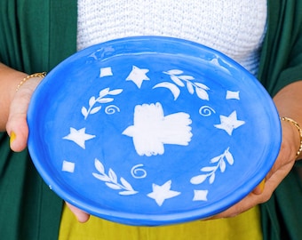 May include: A blue ceramic plate featuring a white bird design and white stars, leaves, and a crescent moon.