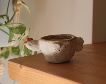 Puede incluir: Una maceta de cerámica con forma de gallina y acabado blanco roto moteado. La maceta tiene una cresta roja, un pico naranja y una abertura redonda para plantas. Está colocada sobre una estantería de madera.