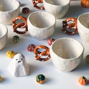 May include: A set of six white ceramic mugs with pretzel handles. The mugs are decorated with small white dots. The mugs are surrounded by small orange and green pumpkins and a white ghost.