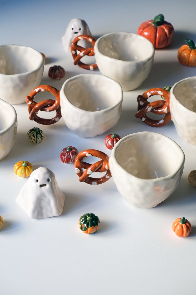 May include: A set of six white ceramic mugs with pretzel handles. The mugs are decorated with small white dots. The mugs are surrounded by small orange and green pumpkins and a white ghost.