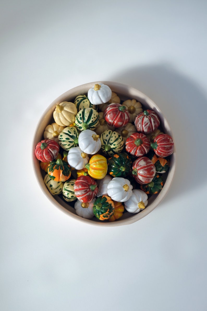 May include: A white bowl filled with a variety of small, colorful gourds. The gourds are painted in shades of green, red, white, and yellow.