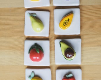Può includere: Otto piastrelle quadrate in ceramica con disegni di frutta. I frutti includono un'arancia, un fico, una pera, una papaya, un pomodoro, un avocado, un limone e una mela. Ogni frutto dai colori vivaci è posto su una piastrella bianca, disposti su una superficie di legno.