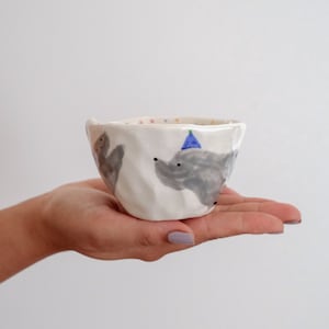 May include: A white ceramic bowl with a textured surface. The bowl is decorated with a gray elephant wearing a blue party hat and colorful dots.